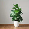 A vibrant green artificial monstera palm plant in a white cylindrical pot displayed on a wooden floor.