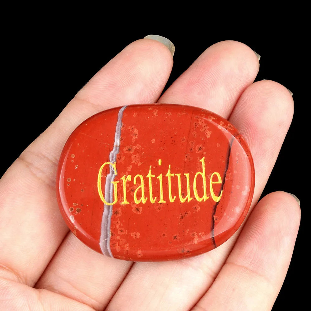 A hand holds a reddish-orange oval crystal with the word Gratitude inscribed on it.