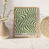 A framed art print featuring a green and cream wavy striped pattern, displayed next to a white vase and a woven sphere.