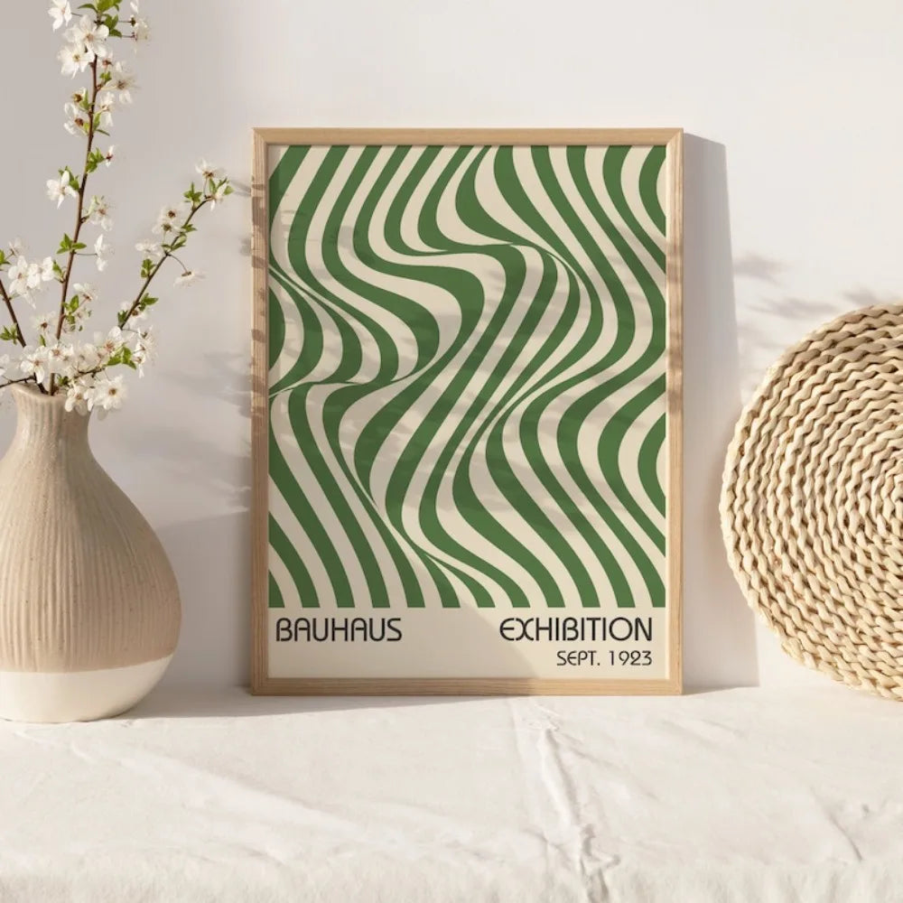 A framed art print featuring a green and cream wavy striped pattern, displayed next to a white vase and a woven sphere.