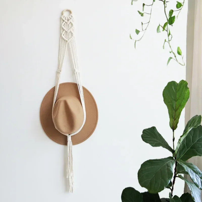A handmade macrame hat organizer with a wooden dowel is displayed as a boho wall hanging against a white wall.