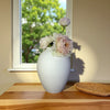 A white ceramic vase holding light pink and white flowers sits on a wooden surface next to a window.