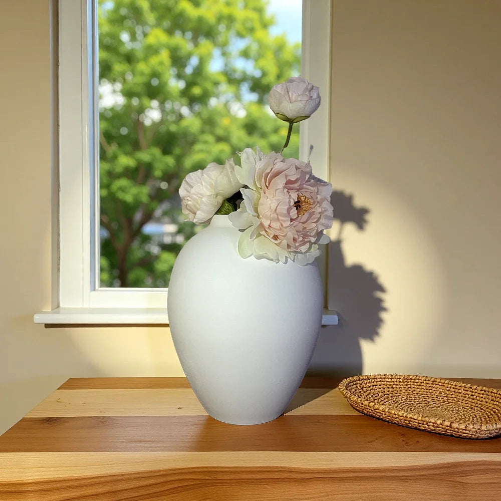A white ceramic vase holding light pink and white flowers sits on a wooden surface next to a window.