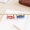 A wooden pencil is placed on a white paper next to a small red crab and a blue crab, used as a desk organizer.