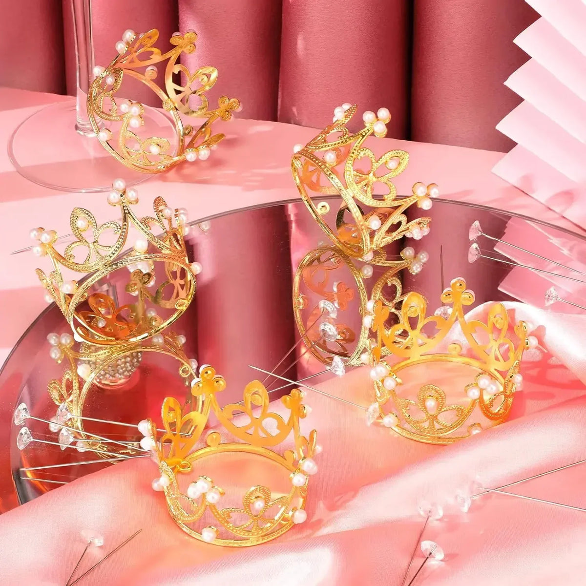 Mini gold and silver crown cake topper decorations displayed on a pink satin background.