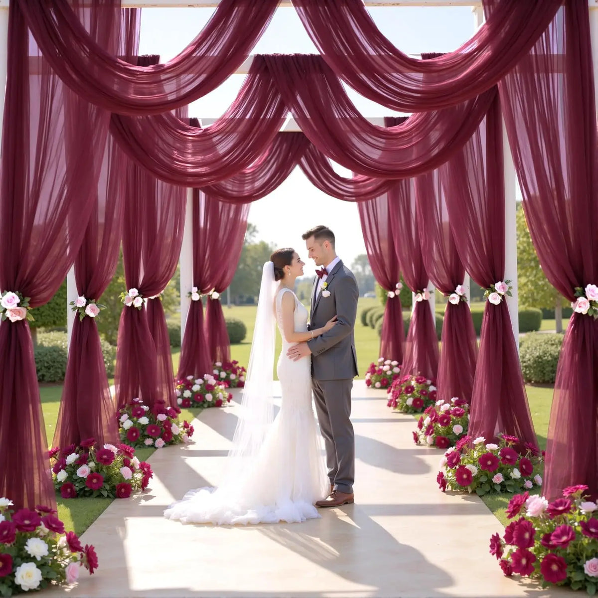 A wedding ceremony setup featuring a deep burgundy tulle draped backdrop and aisle decorated with pink and white flowers.