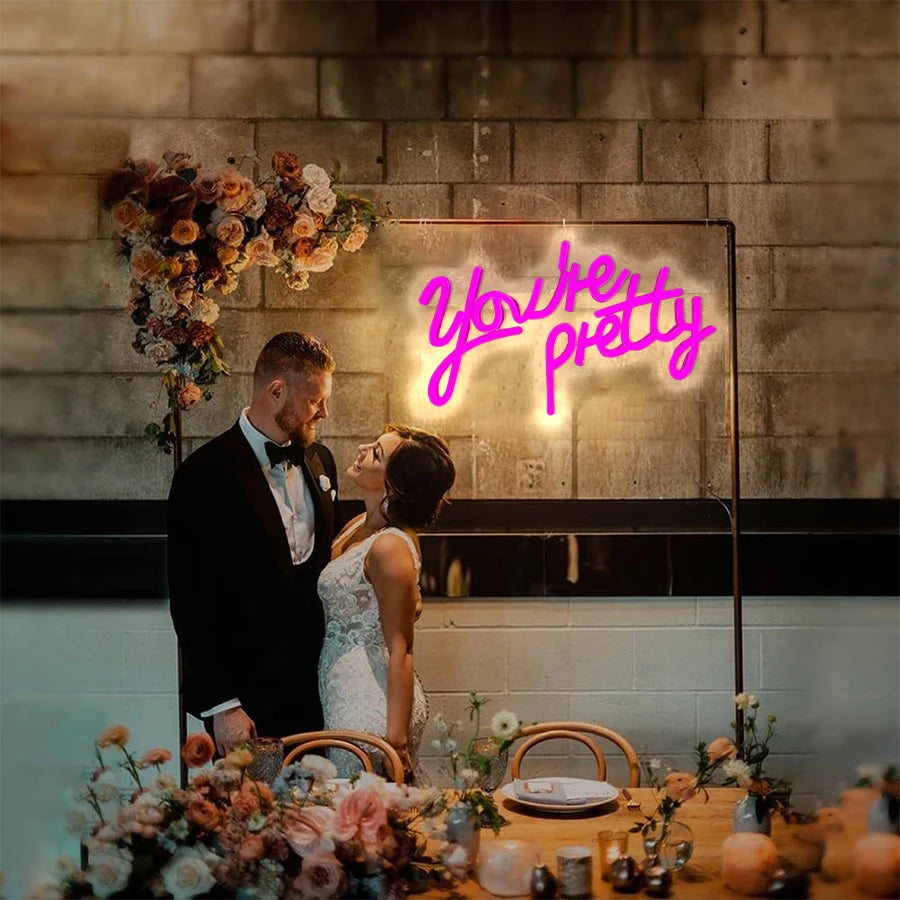 A couple is looking at a neon sign that reads Youre pretty mounted on a brick wall.