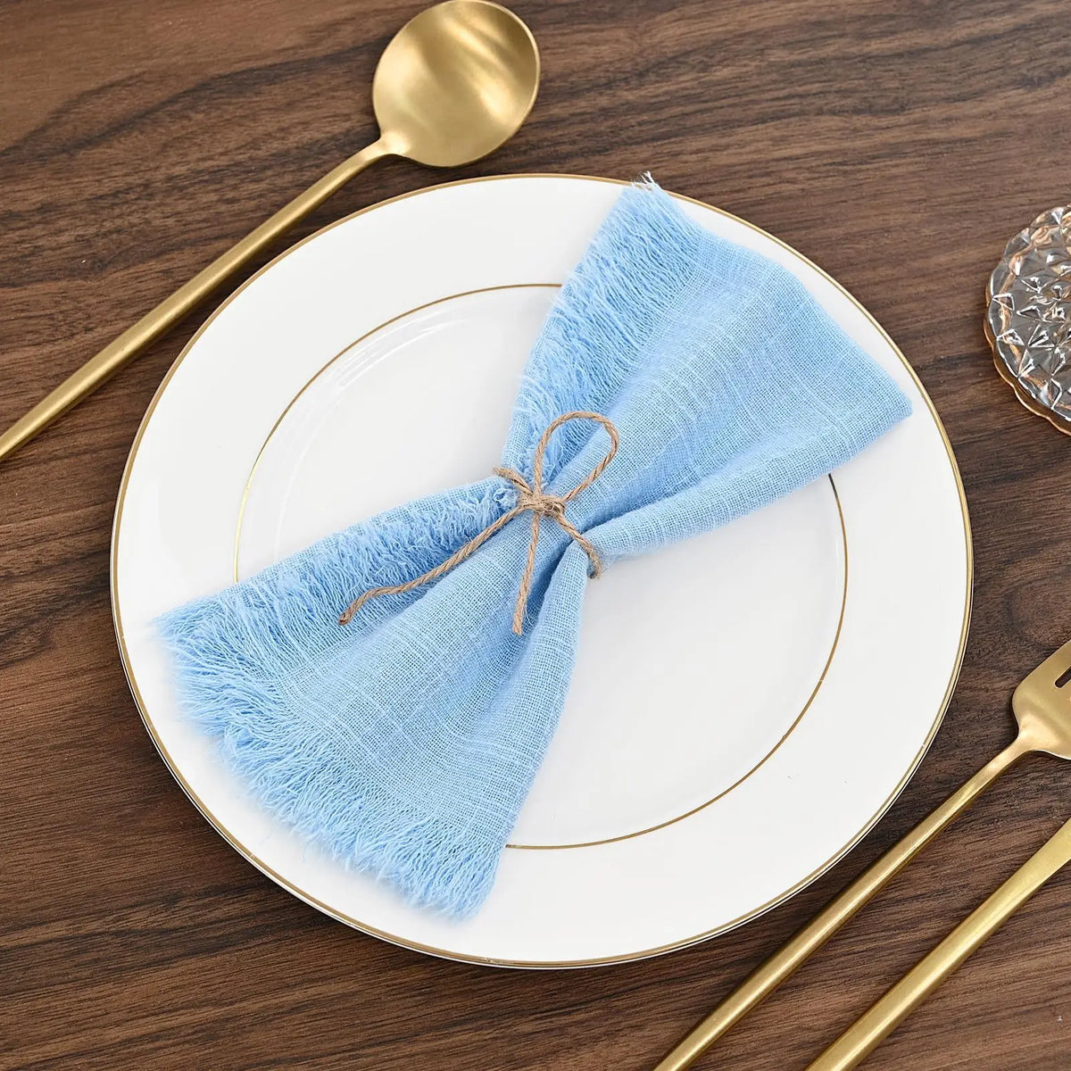 A set of ten light blue fringed cotton napkins placed on a white plate with gold rim and gold cutlery.