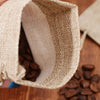 A close-up view of a natural burlap drawstring bag partially open, showing dark roasted coffee beans inside.