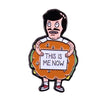 An enamel pin featuring a cartoon man with a surprised expression holding a sign that reads This is me now