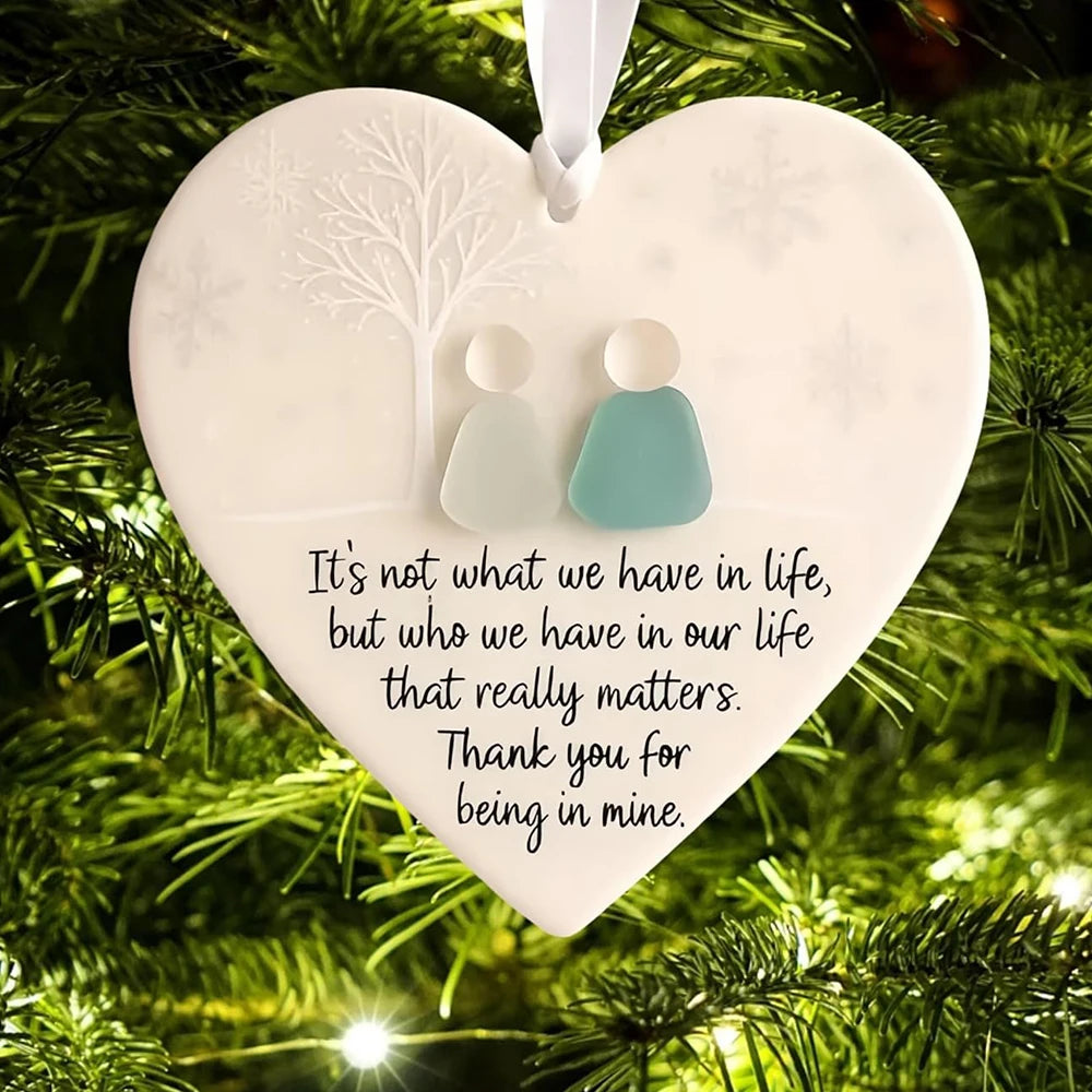 A white heart-shaped ornament with a frosted tree silhouette and two small figures, set against a green pine branch backgroun