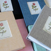 A collection of photo albums with linen covers and floral designs, showing various shades of pink, blue, and green.