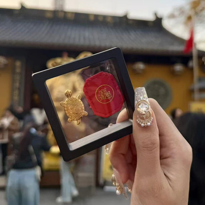 A hand holds a gold tortoise figurine for wealth luck home decor against a traditional building background.
