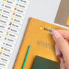 A hand is placing a yellow name sticker reading Martin onto an orange spiral notebook page filled with similar stickers.