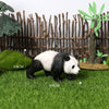 A realistic panda figurine standing on green grass with a bamboo fence and small plants in the background.