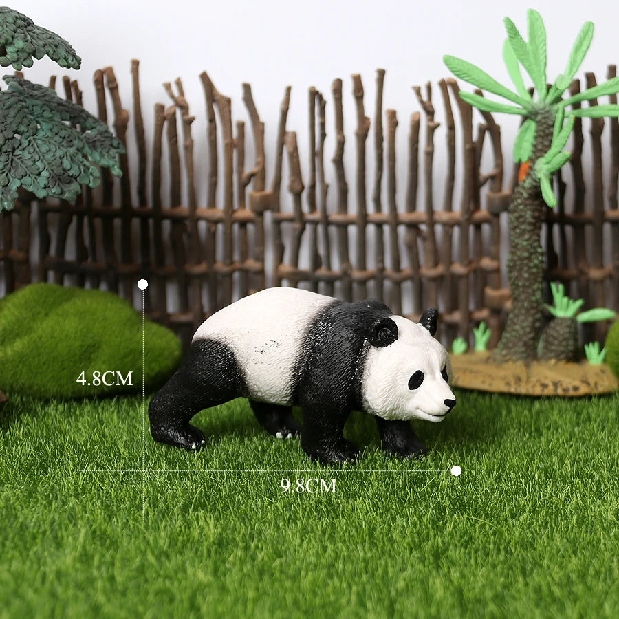 A realistic panda figurine standing on green grass with a bamboo fence and small plants in the background.