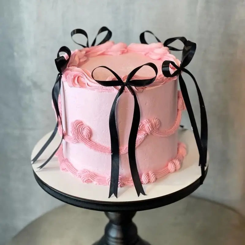 A two-tiered pink cake decorated with ruffled buttercream and black ribbon bows.