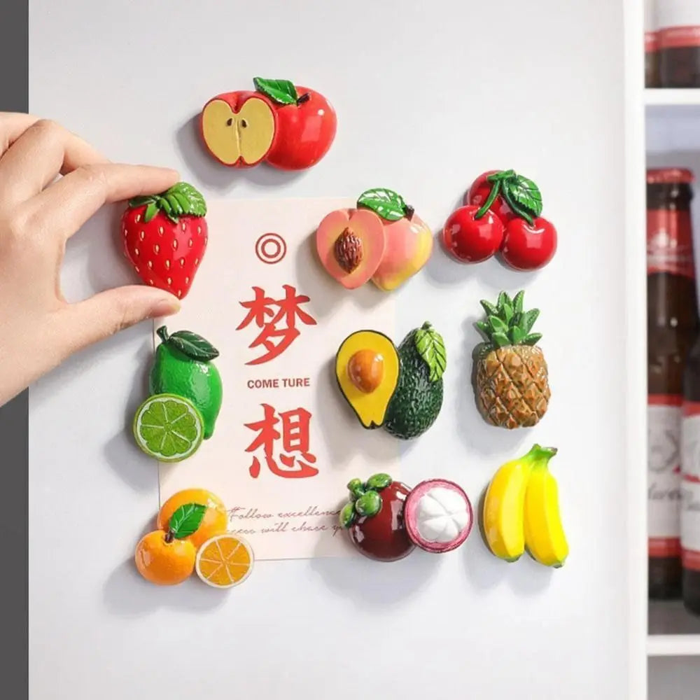 A hand places various colorful fresh fruits like apples strawberries and pineapples onto a white board for fridge magnets.