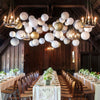 A long wooden dining table set for an event with white linens and decorative paper lanterns hanging above.