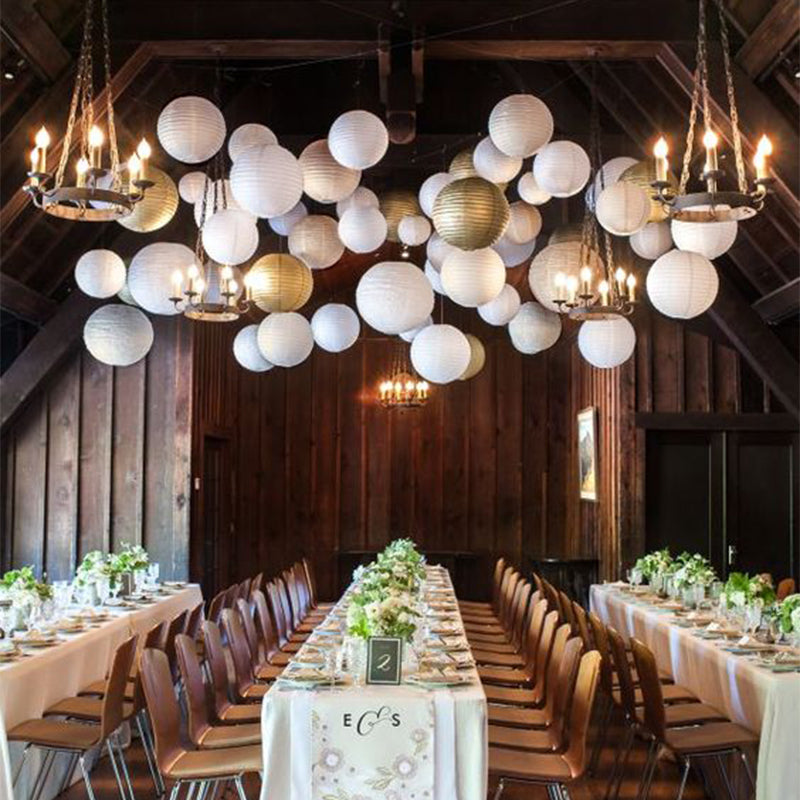 A long wooden dining table set for an event with white linens and decorative paper lanterns hanging above.