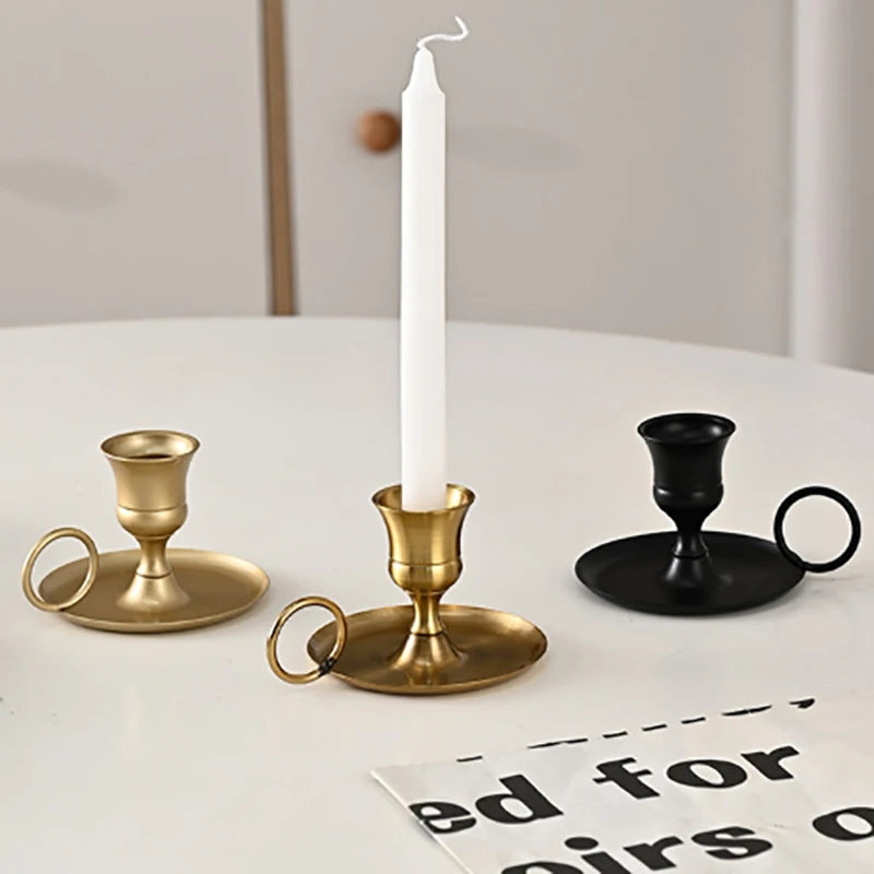 A tall white candle in a brass metal holder with a handle sits on a white table next to a black candle holder.