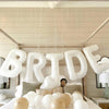 A display of white letter foil balloons spelling BRIDE hanging from a wooden frame against a light background.