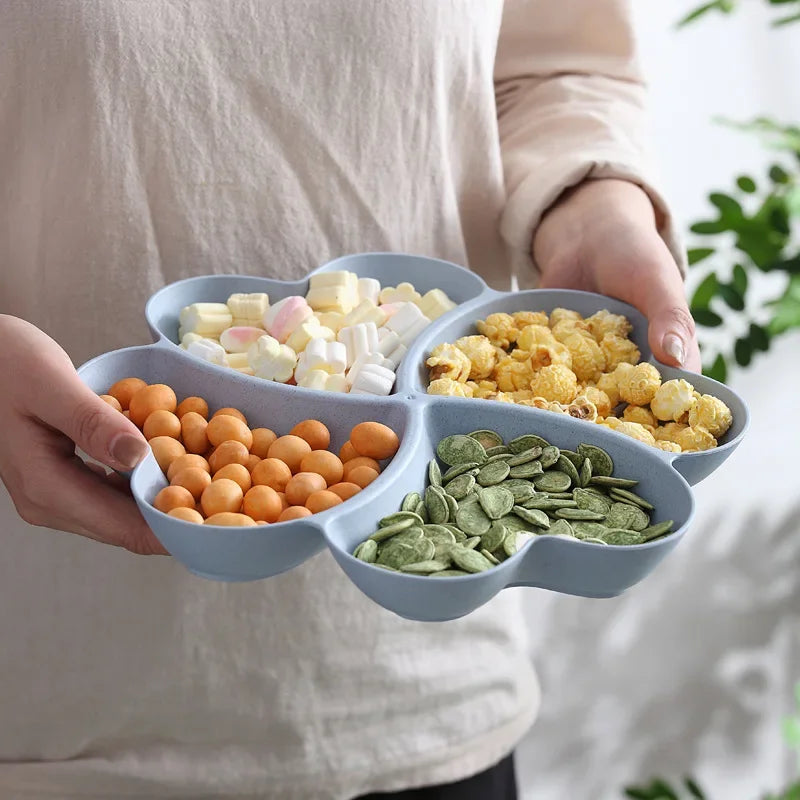 A hand holds a heart-shaped four-compartment snack tray filled with various seeds and nuts.