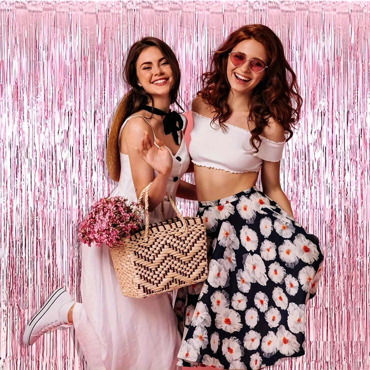 Two women posing against a pink metallic foil tassel backdrop wearing white and floral patterned clothing