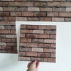 A close-up view of a rectangular brown brick pattern wall sticker being held up against a white background.