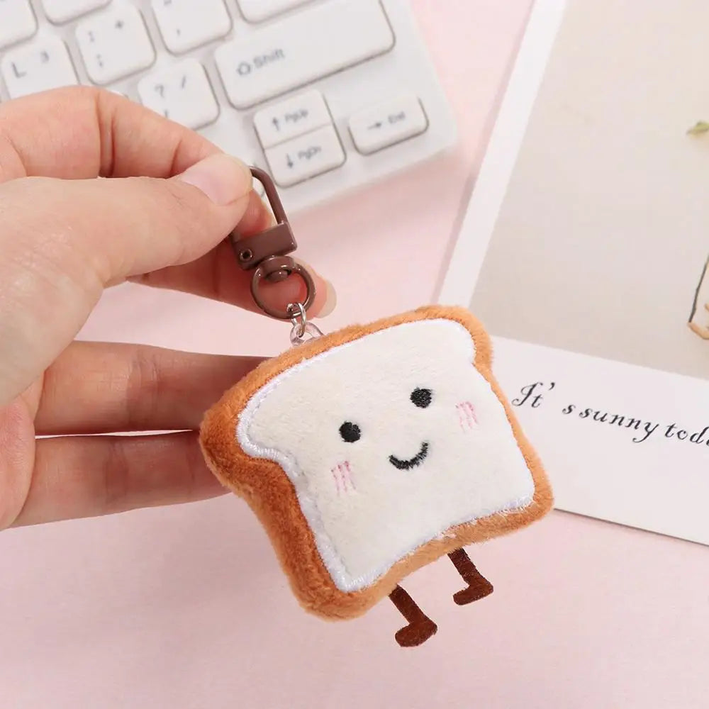 A fluffy toast bread plush charm with brown edges and small pink eyes is being held by a hand against a white background.