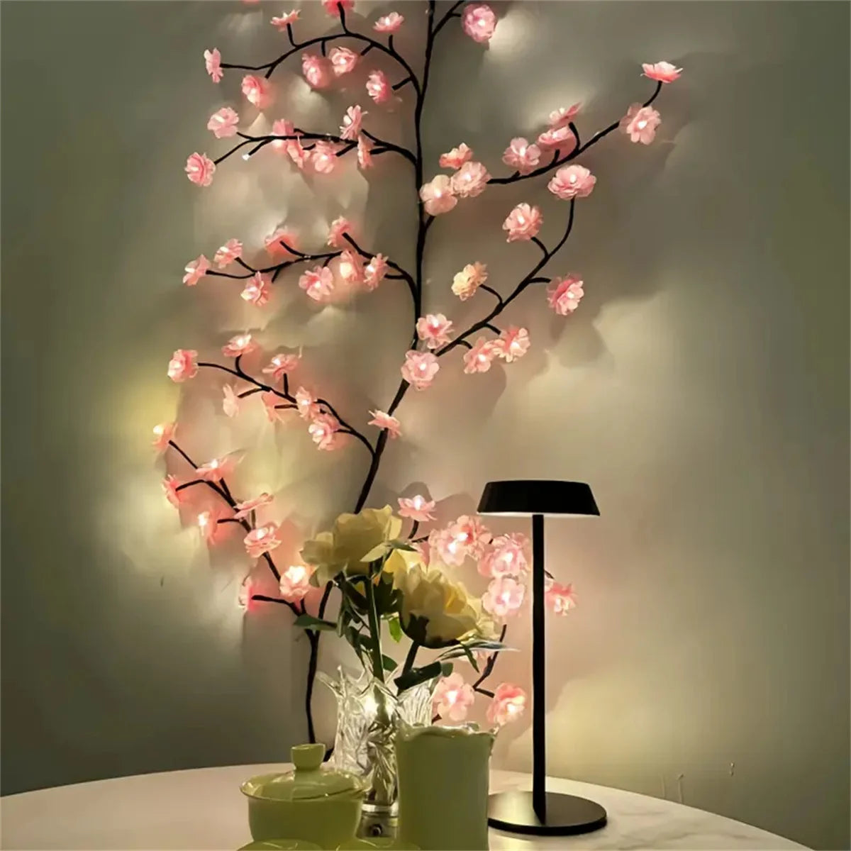 A branch of pink LED rattan string lights decorated with small flowers and a white rose in a vase on a table.