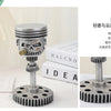 A silver mechanical piston skull sculpture with a gear base and piston rod, displayed on a white surface.