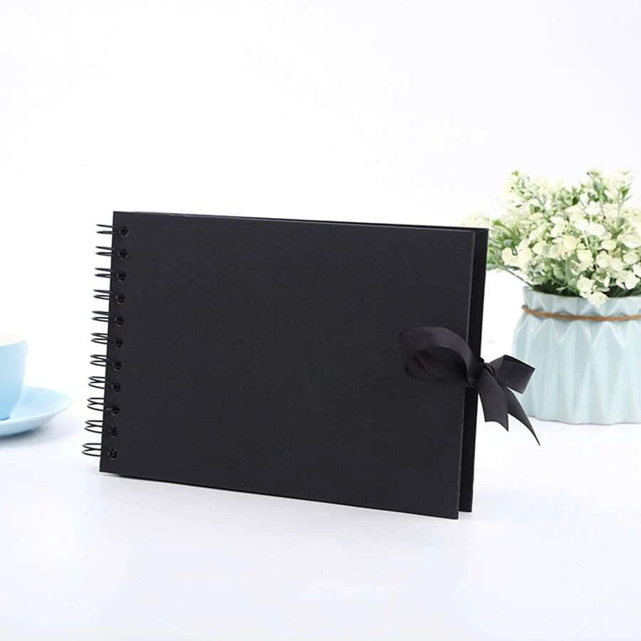 Un cuaderno encuadernado en espiral negro con una cinta negra atada en la cubierta junto a una planta de flores blancas.