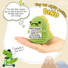 A handmade crochet dinosaur doll is held in a hand with an inspirational message about staying strong and having courage.