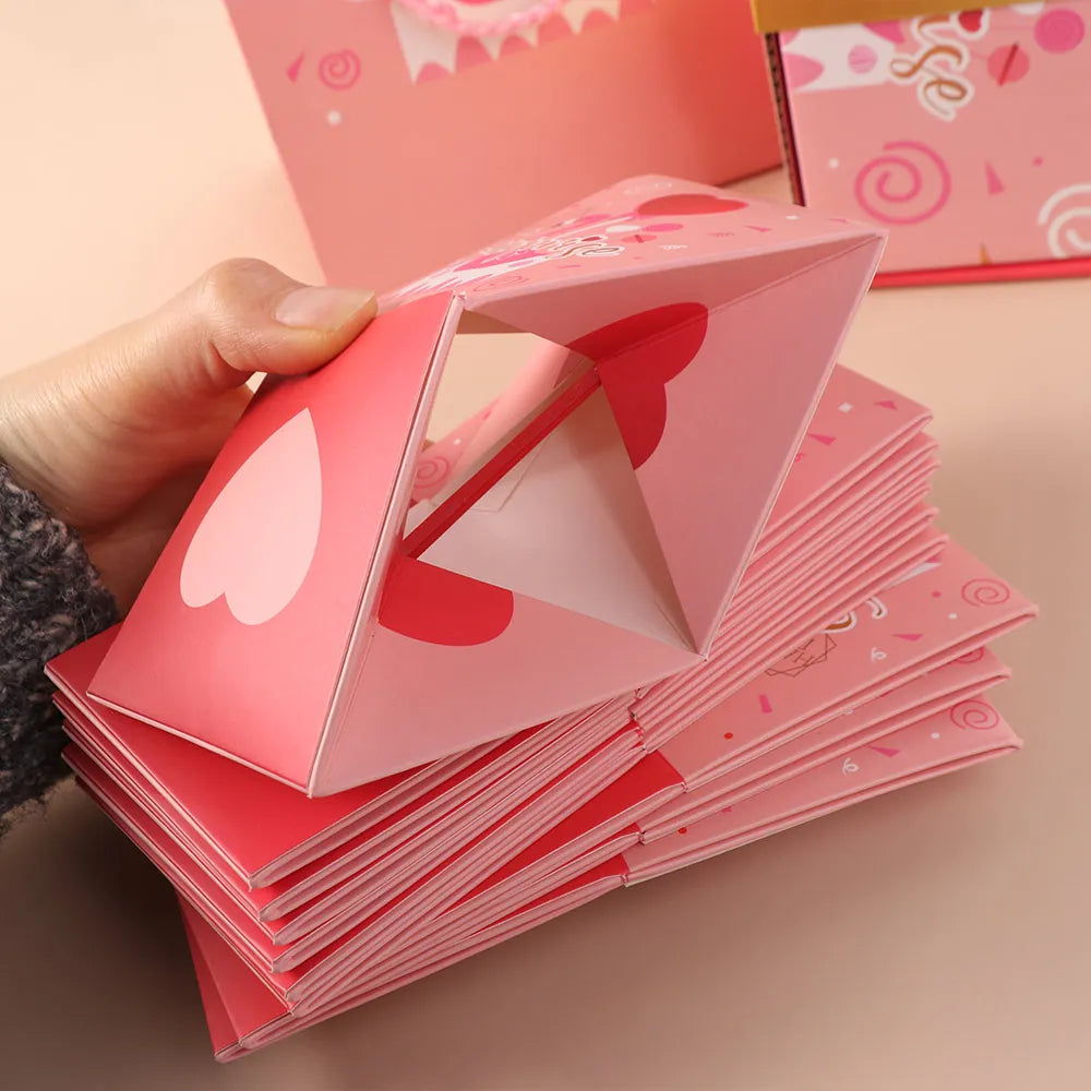 A hand holds a pink luxury pop-up surprise gift box, showing the folded cards and decorative elements.