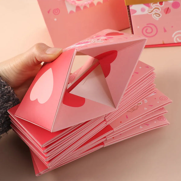 A hand holds a pink luxury pop-up surprise gift box, showing the folded cards and decorative elements.