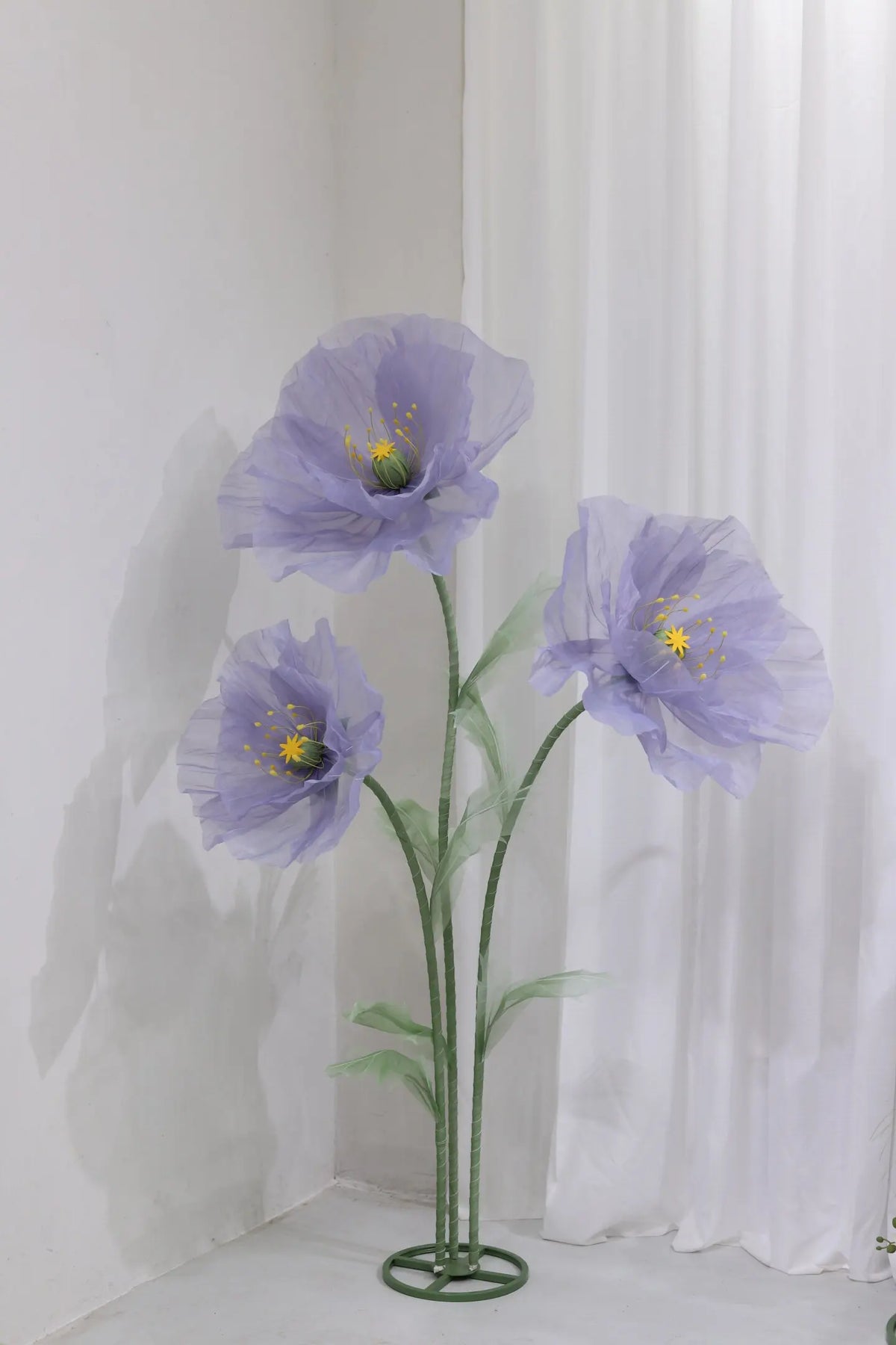 A large artificial silk purple poppy flower with a green stand displayed against a white wall.