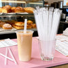 A glass jar filled with clear plastic drinking straws is displayed next to a glass of light orange beverage.