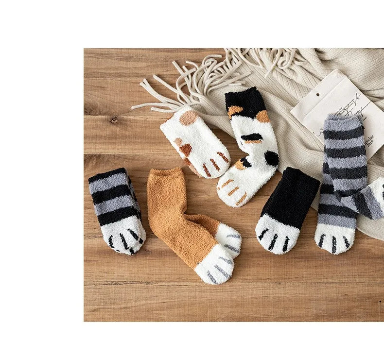 A set of cozy cat paw print plush socks in black, white, and tan colors displayed on a wooden surface.