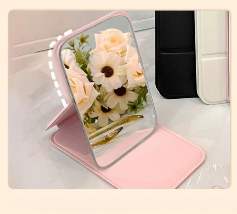 A pink portable folding HD makeup mirror with a floral design resting on a white surface.