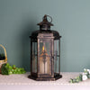 A vintage Nordic iron candle lantern with glass panes sits on a white surface next to green grapes and foliage.