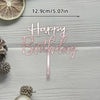 A pink acrylic birthday sign spelling out Happy Birthday with a stick, shown next to a slice of citrus and pinecones.