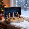 A silver tin holds a nativity scene figurine set featuring figures like Mary, Joseph, and animals on straw bedding.