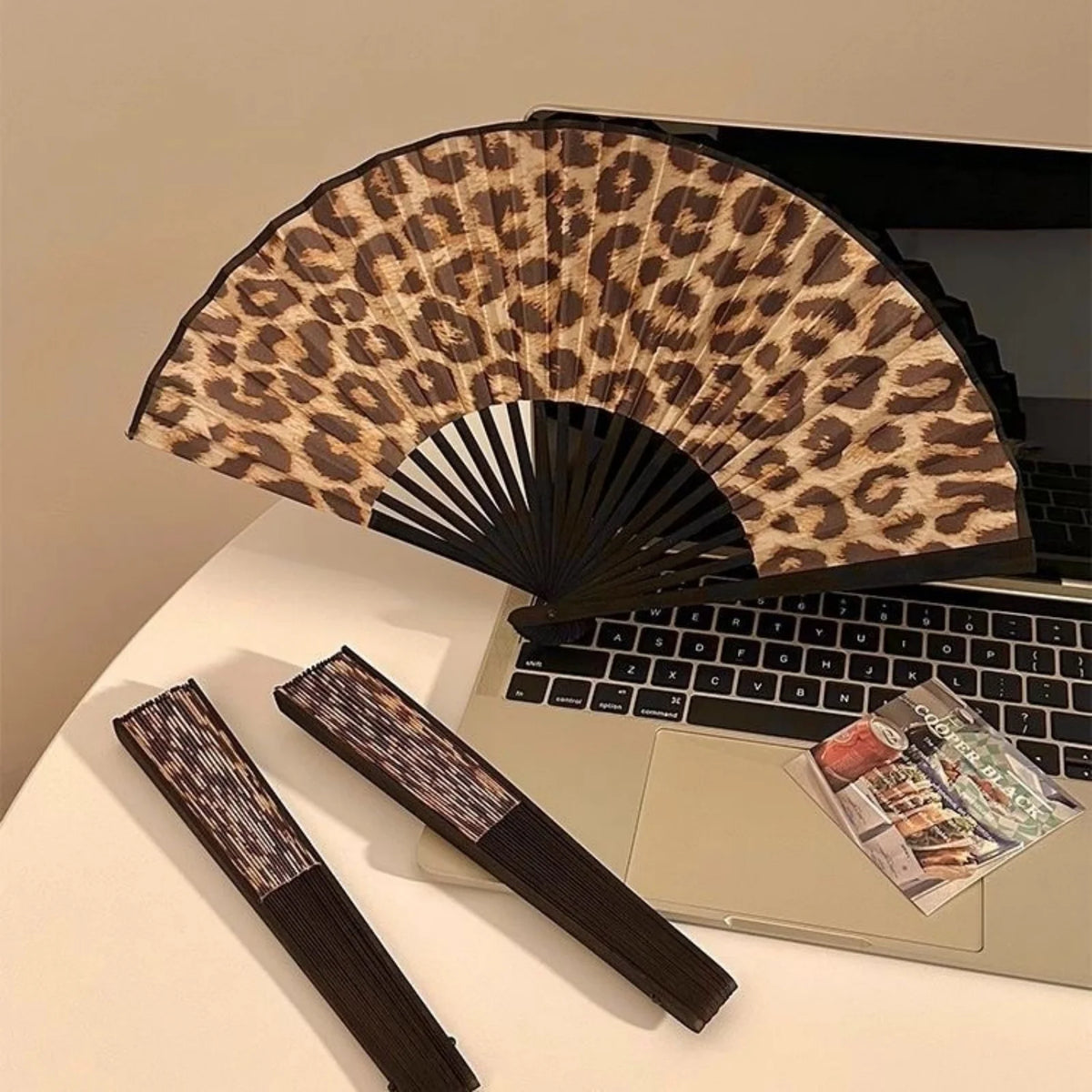 A leopard print folding hand fan with a black frame resting on a laptop.