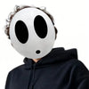 A white, smooth mask resembling a stylized face with large black eye holes and a small mouth, worn by a person in a dark hood