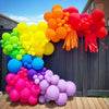 A colorful archway made of latex balloons in shades of orange, yellow, pink, and red against a dark wooden fence.