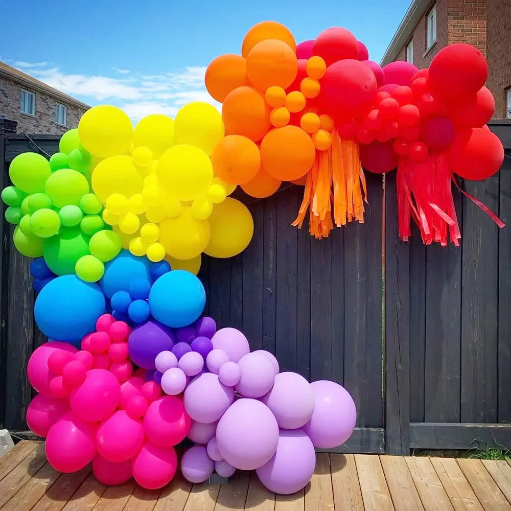 A colorful archway made of latex balloons in shades of orange, yellow, pink, and red against a dark wooden fence.