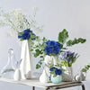 A still life arrangement featuring blue roses and greenery in white and light green ceramic vases.