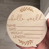 A round wooden sign for a newborn keepsake with lines for name, date, weight, and length, surrounded by a laurel wreath desig