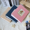 A pink linen cover photo album with a floral design is displayed next to several books on a white textured surface.
